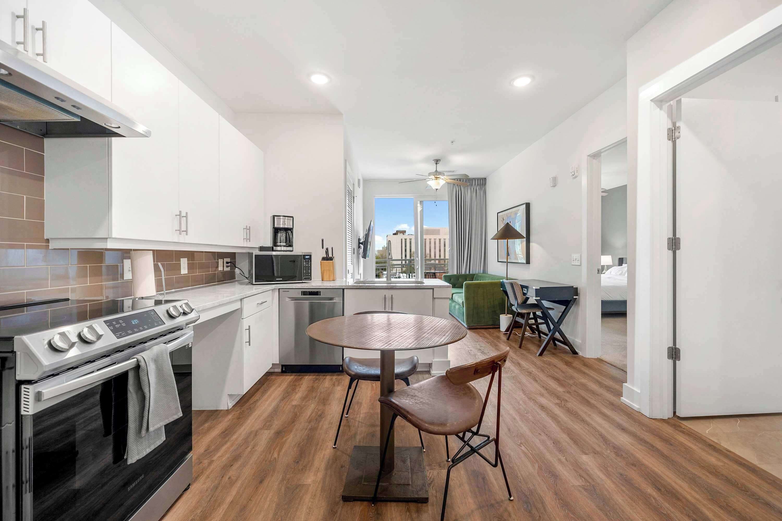 A modern kitchen with a built-in oven, microwave, and coffee maker. There is a dining table with two chairs and a view of the city through large windows.