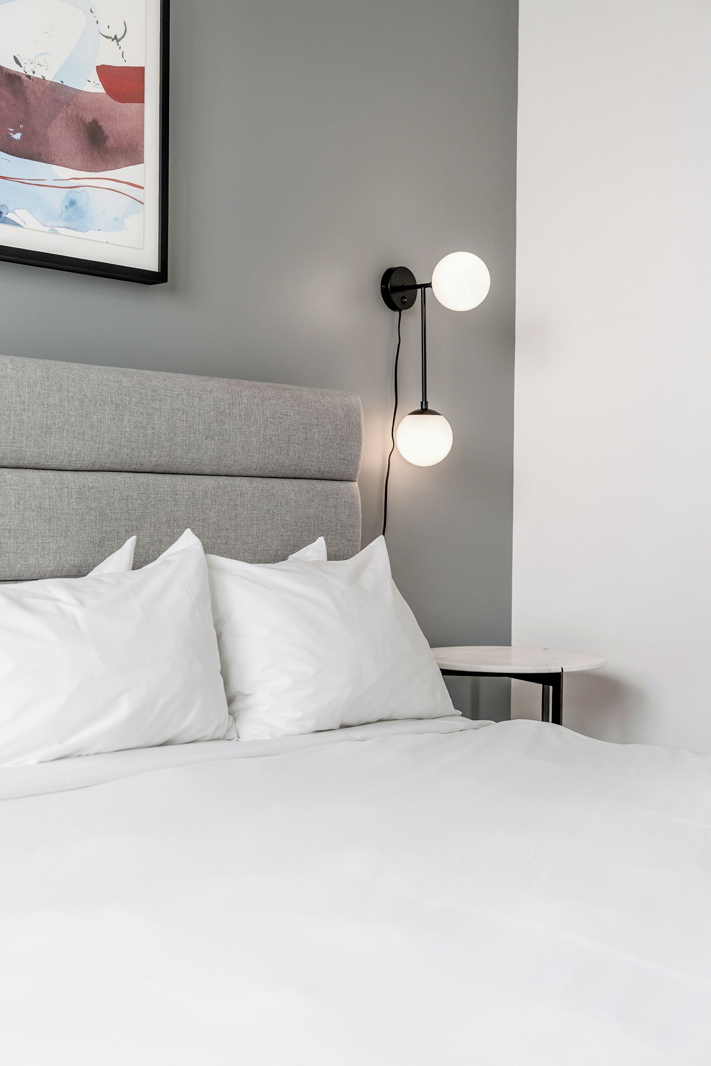 Una cama bien hecha con ropa blanca y almohadas, un cabecero tapizado en gris y una lámpara de pared con dos bombillas redondas.