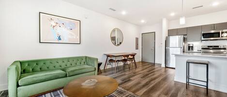 A modern living room with a green sofa, a round coffee table, and a dining area with a table and chairs. A kitchen with a refrigerator, microwave, and stove is visible in the background.