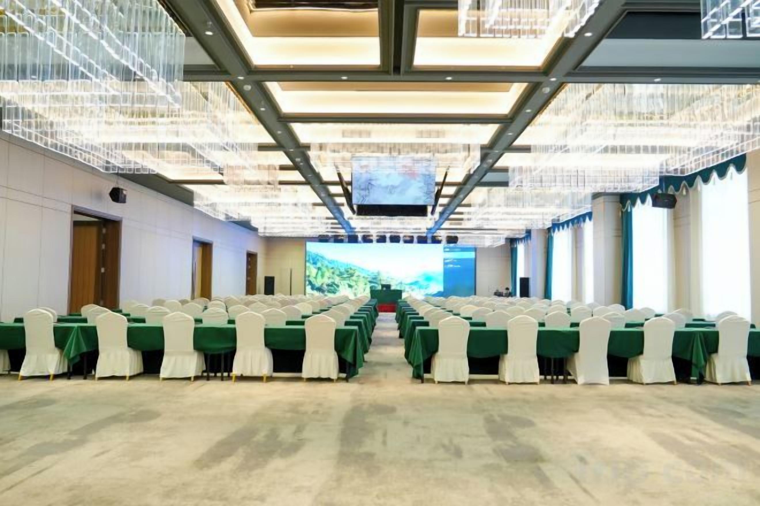 A conference room with rows of tables and chairs, a large screen at the front, and a modern ceiling design.
