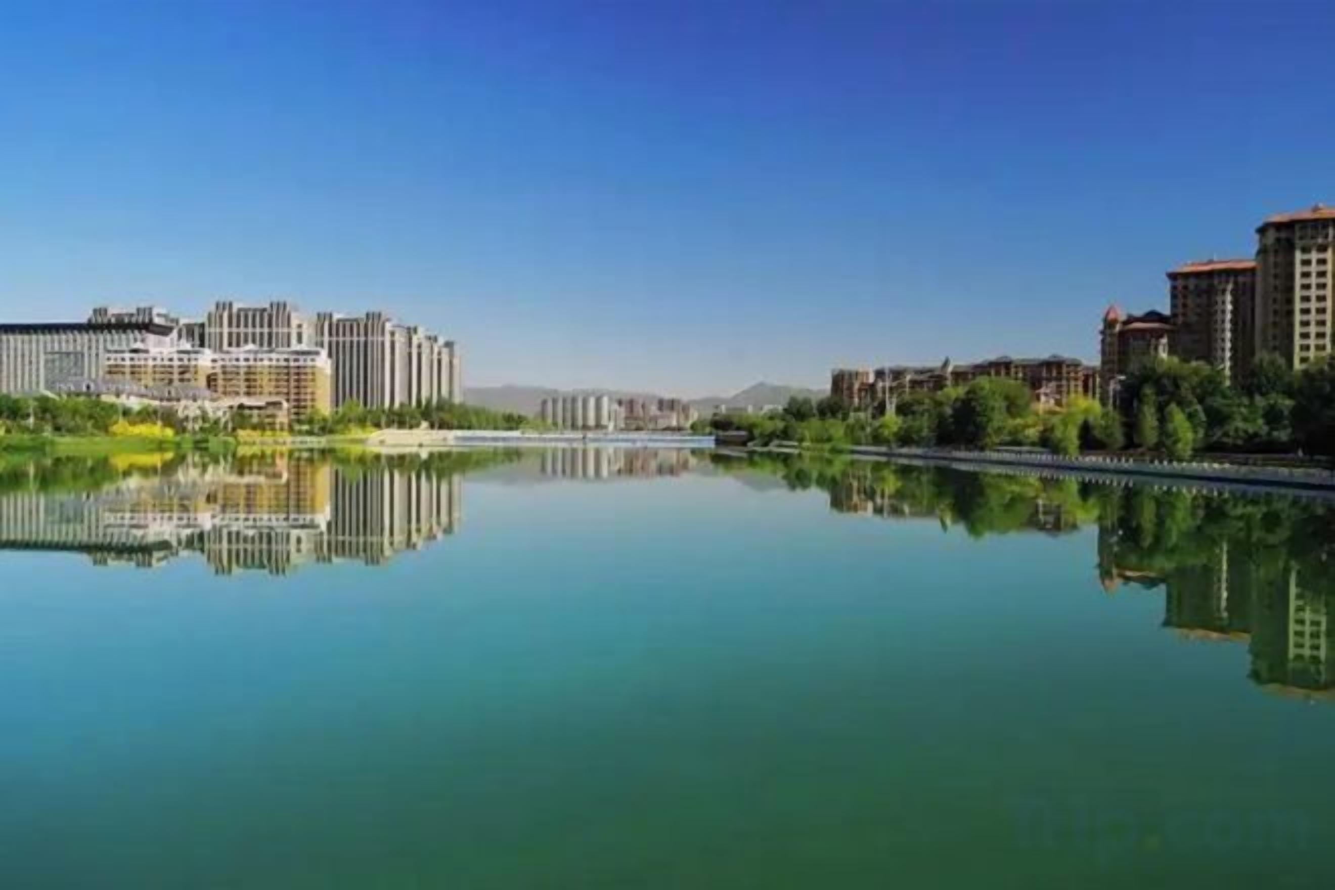 A calm river reflecting buildings, a clear sky, and a distant mountain range.