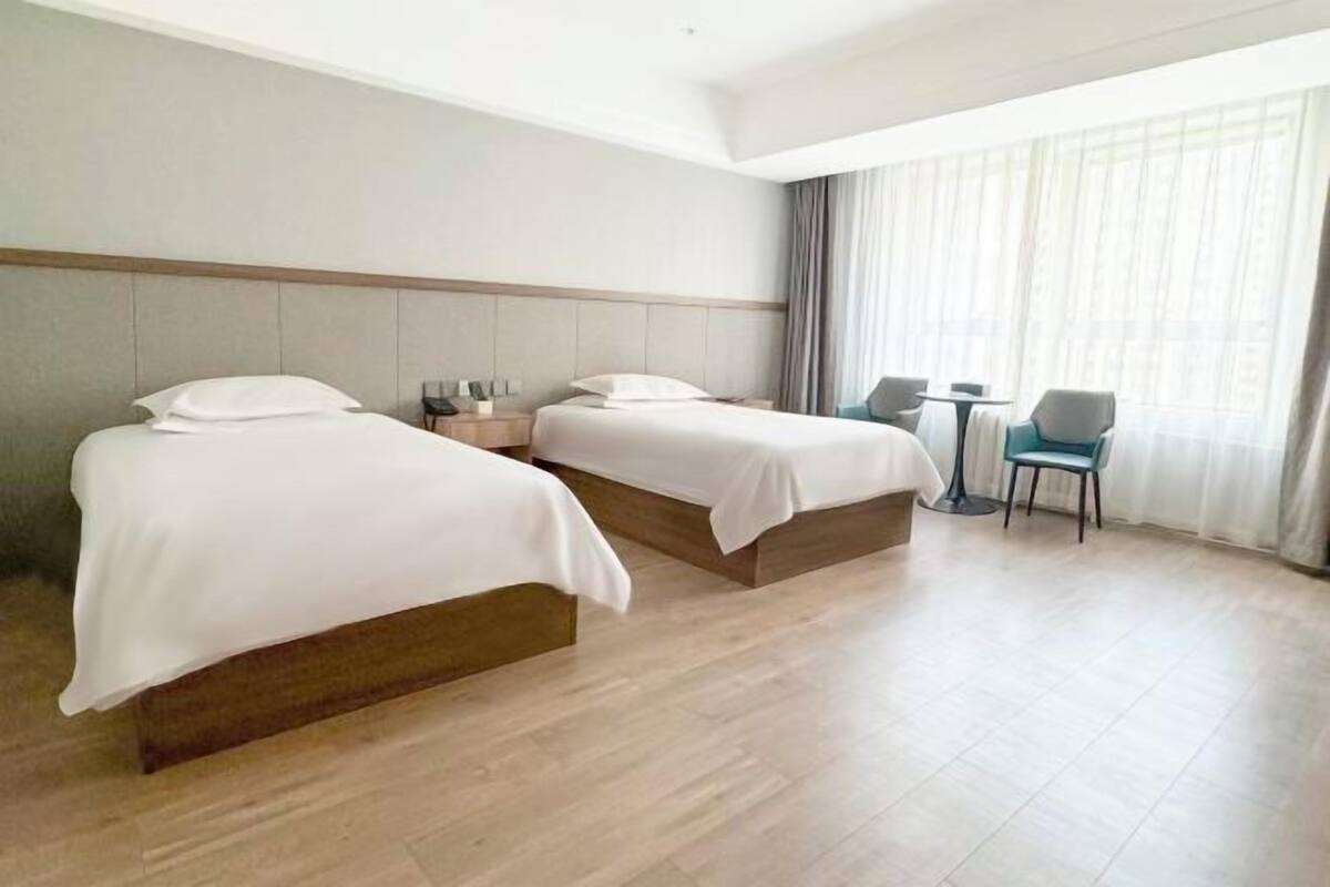 Une chambre d’hôtel avec deux lits, une petite table, des chaises et une grande fenêtre avec des rideaux.
