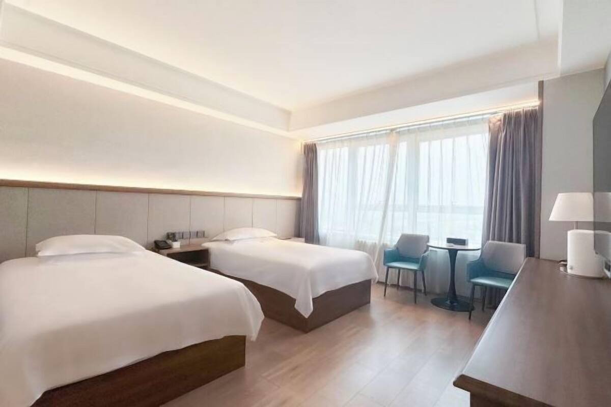 Une chambre d’hôtel avec deux lits, un bureau, une chaise, une petite table et une fenêtre avec des rideaux.