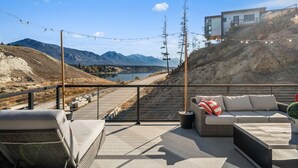 A terrace with outdoor seating, a view of a lake, mountains, and a building on a hillside. - Salt & Stone | Lake/Private Beach | Mountain Views (Invermere)