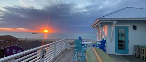 A wooden deck with a table and chairs, overlooking a beach with a sunset.