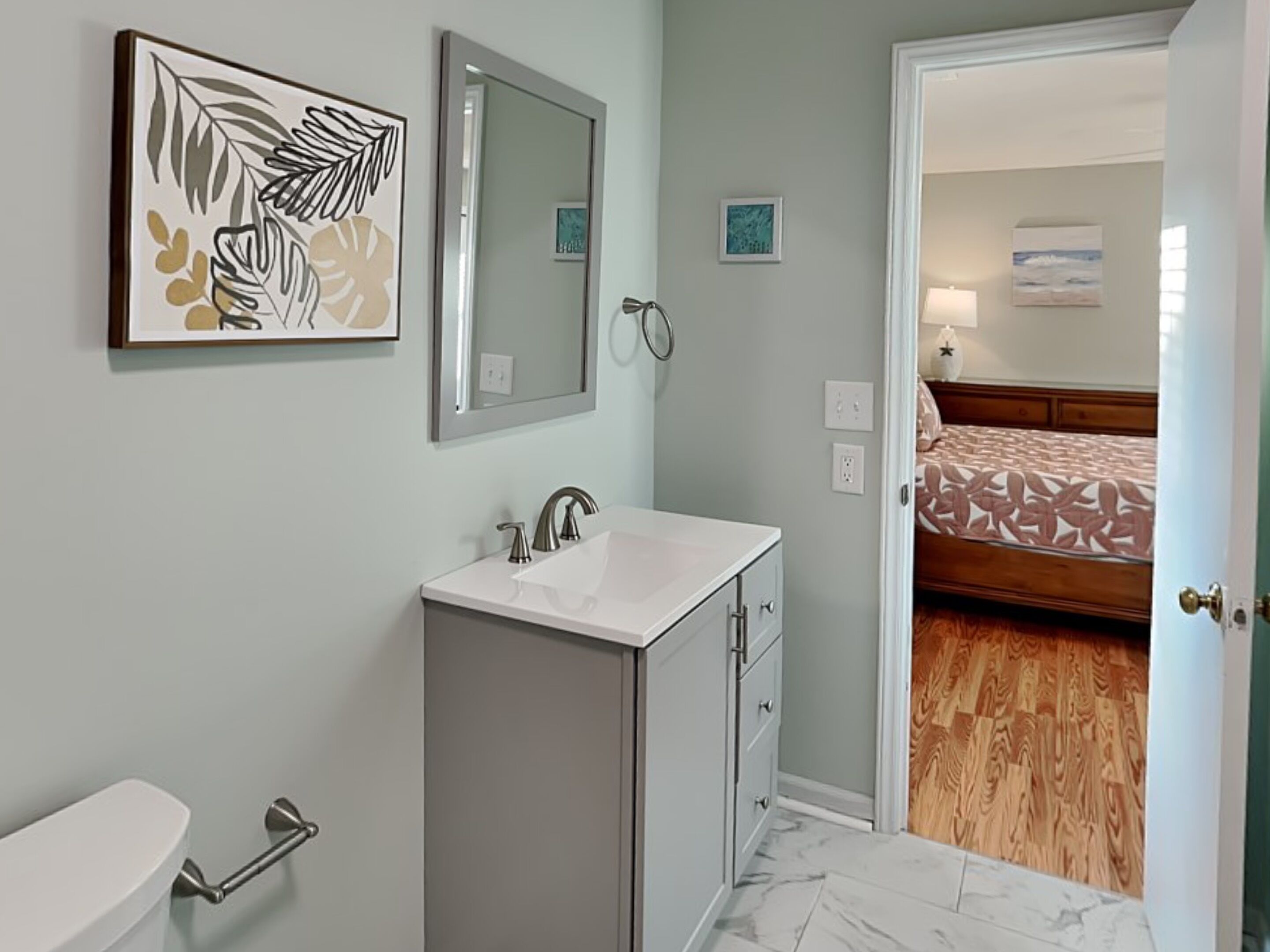 Baño con lavabo, espejo y retrete. Una puerta da acceso a un dormitorio con cama y suelo de madera.