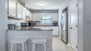 A modern kitchen with white cabinets, a stainless steel refrigerator, a microwave, and a coffee maker. There are two white barstools and a window with blinds. - Carolina Cottage by Carolina Retreats (Carolina Beach)