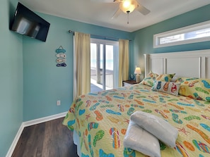 A bedroom with a bed, a ceiling fan, a TV mounted on the wall, and a window with curtains. - Shore Thing by Topsail Realty (Holly Ridge)