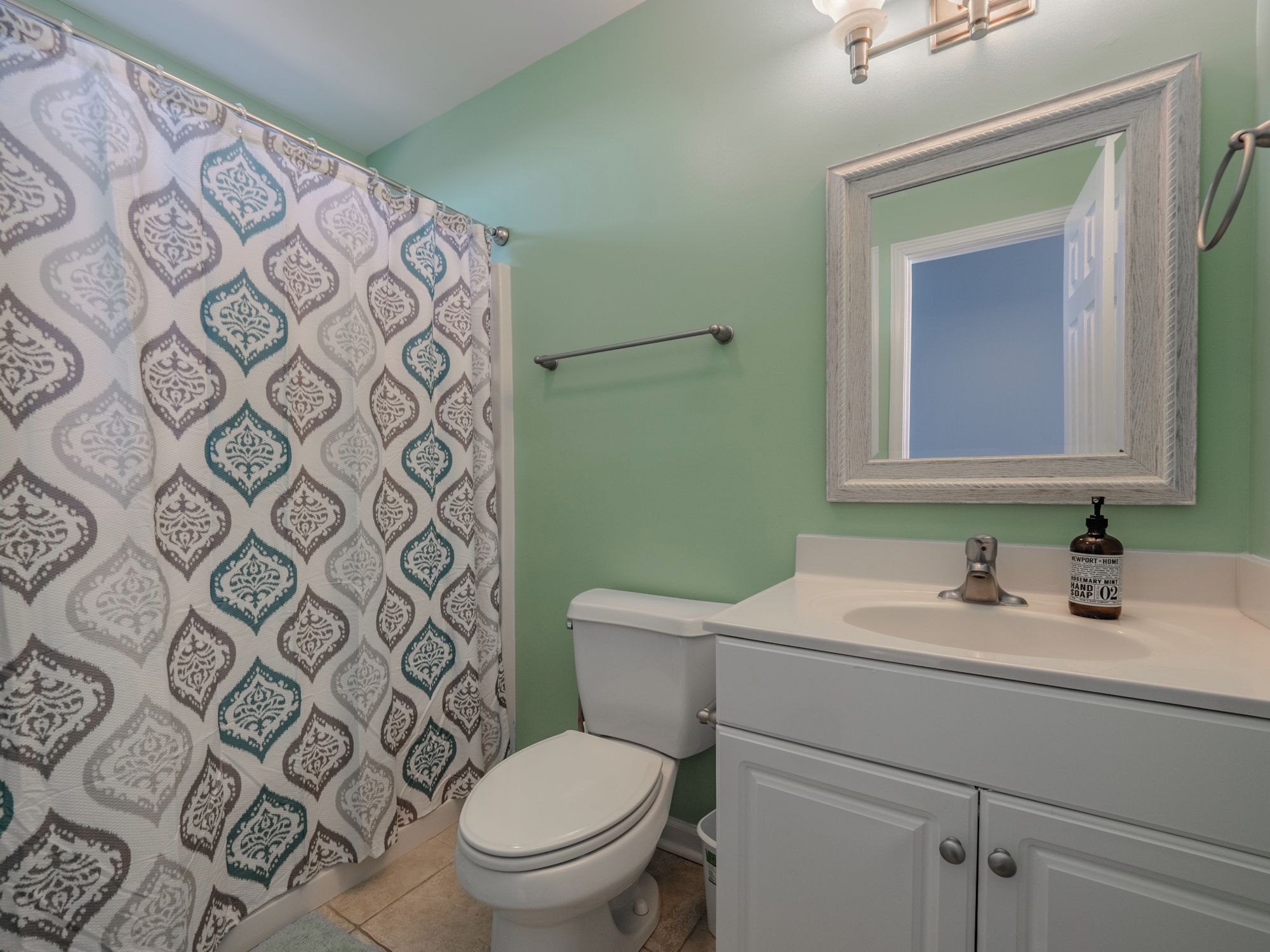 A bathroom with a patterned shower curtain, a toilet, a sink with a soap dispenser, and a mirror above the sink.
