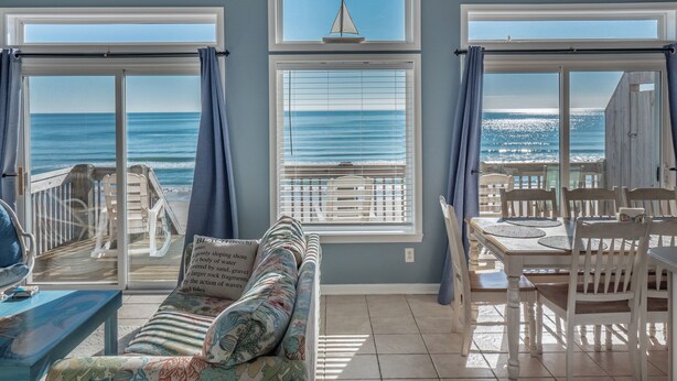 A room with a view of the ocean, a sailboat on the window, a sofa, and a dining table.