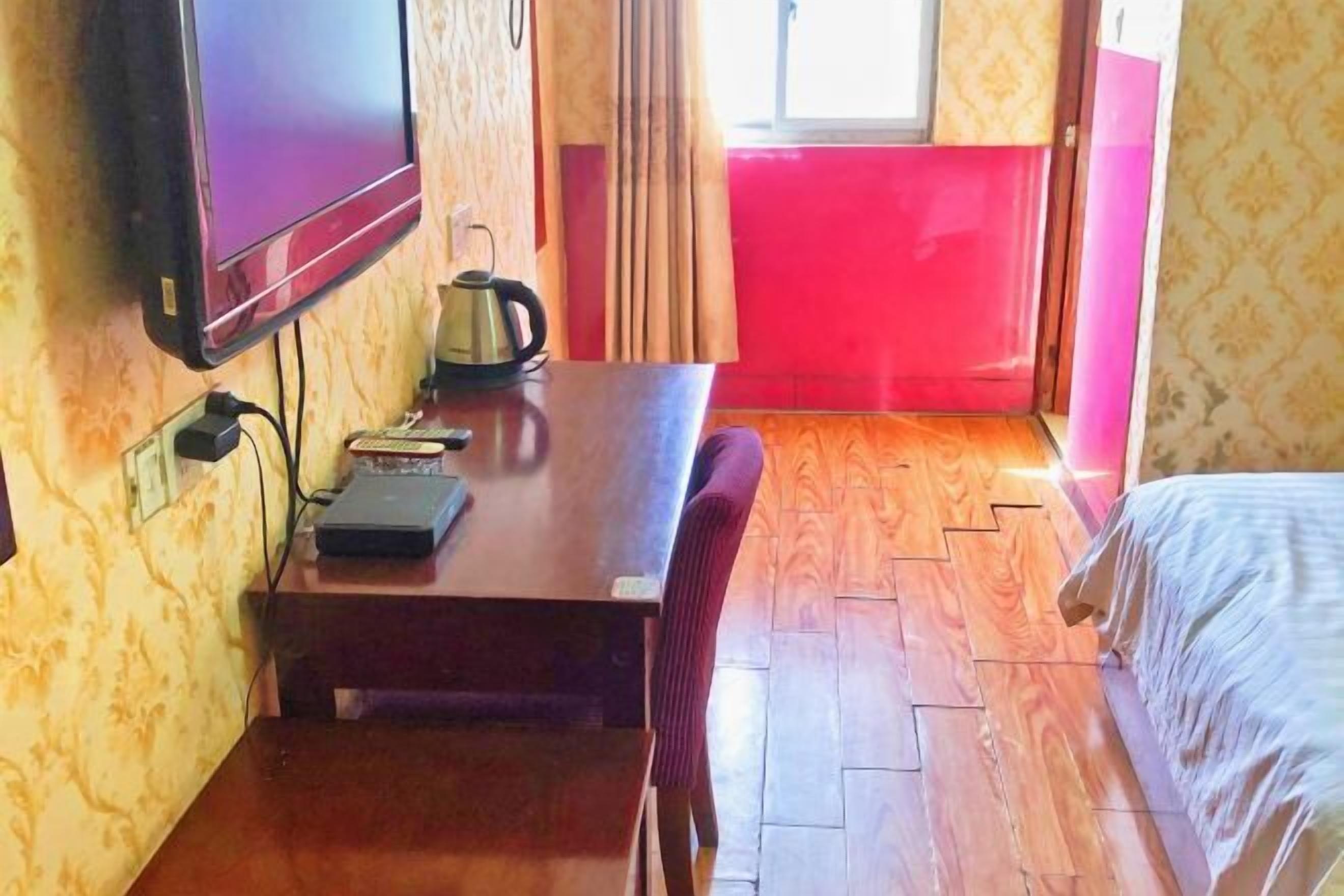 A room with a wooden table, a television mounted on the wall, a kettle, and a bed with a white cover.