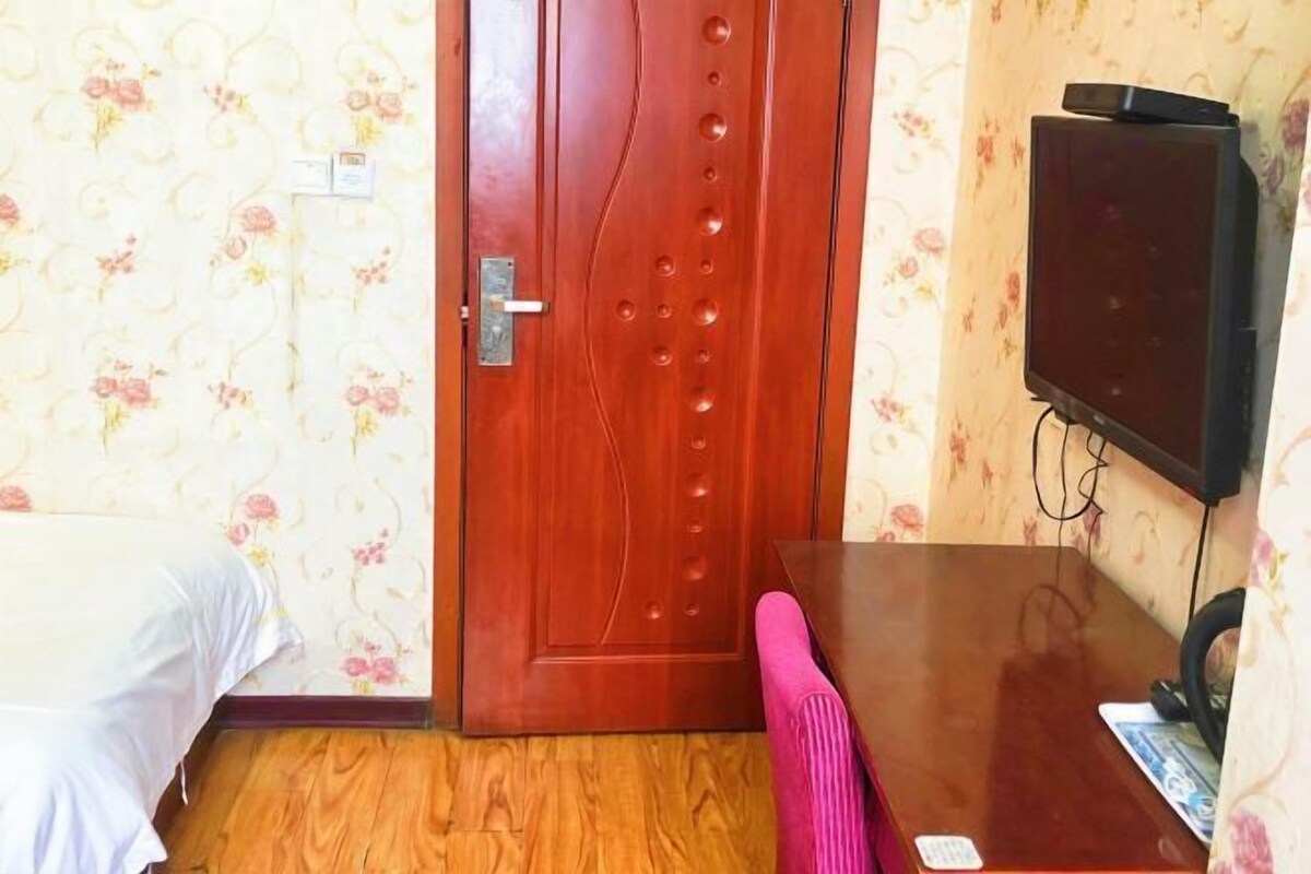 Une pièce avec une porte en bois, un lit recouvert d’une couverture blanche, une table en bois avec une chaise rose, et un téléviseur fixé au mur.