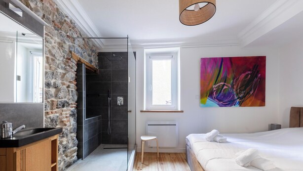 Ein modernes Schlafzimmer mit Steinwand, einem großen Bett, einer Dusche und einem farbenfrohen abstrakten Gemälde.