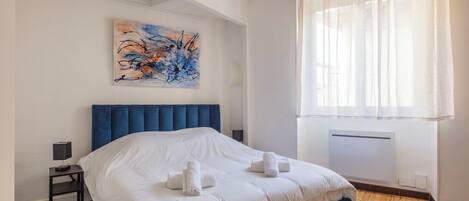 A modern bedroom with a large bed, a nightstand, a wall-mounted abstract painting, and a window with sheer curtains.