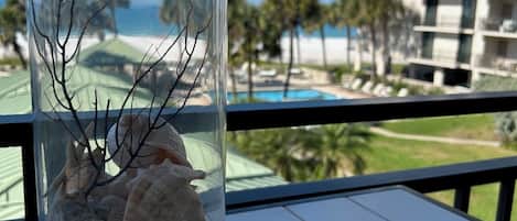 En genomskinlig glasvas med snäckskal och grenar står på en balkong med utsikt över en pool och en hotellbyggnad.