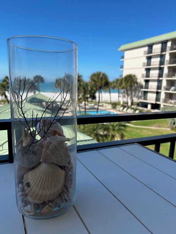 A clear glass vase with seashells and branches on a balcony overlooking a pool and hotel building.