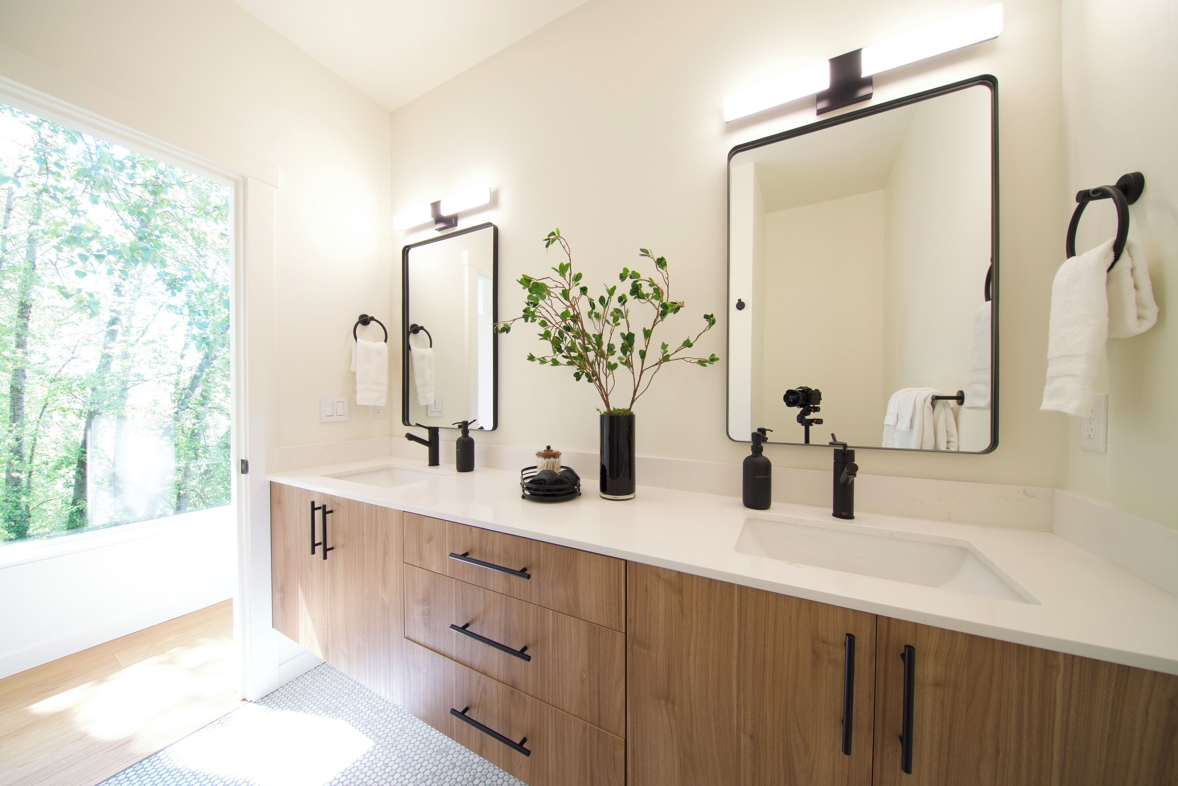 Un baño moderno con un ventanal amplio, muebles de madera, encimera blanca y dos espejos.