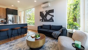 Una sala de estar moderna con sofá, una mesa de centro y taburetes de bar. Se ve un ventanal con vistas a zonas verdes.