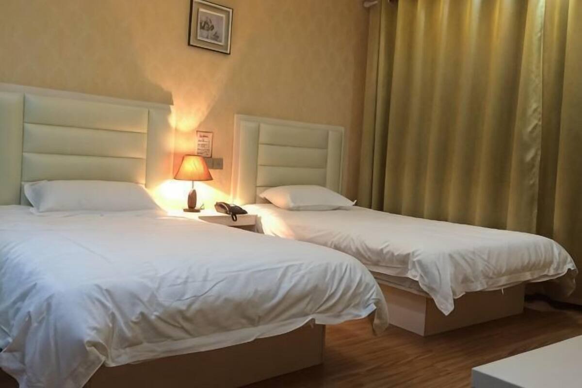 Twee enkele bedden met witte lakens, een nachtkastje met een lamp, en een lijst met een afbeelding aan de muur.