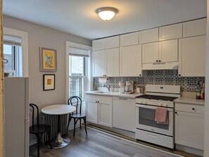 A kitchen with white cabinets, a round dining table, a black chair, and a window with blinds.