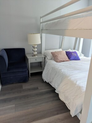 A bunk bed with white bedding, a bedside table with a lamp, and a dark blue chair.