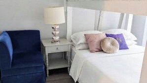 Una cama litera con cabecera blanca, una mesita de noche con lámpara, una silla azul y una cama con varias almohadas.
