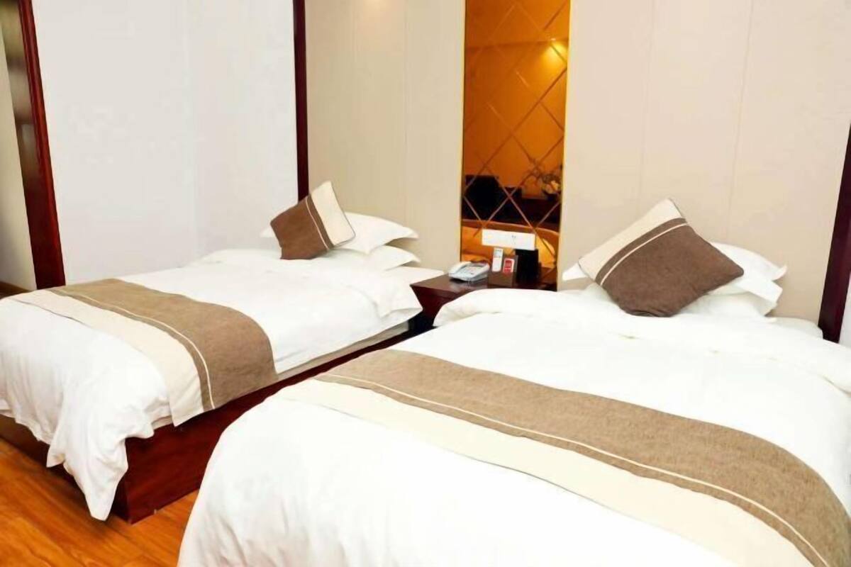A hotel room with two beds, each with white bedding and brown accents, a wooden headboard, a nightstand with a lamp, and a mirror.