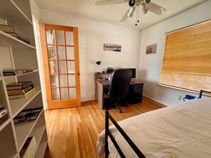 Une chambre à coucher comprenant un lit, un bureau avec une chaise, une bibliothèque, une fenêtre avec des stores et un ventilateur de plafond.
