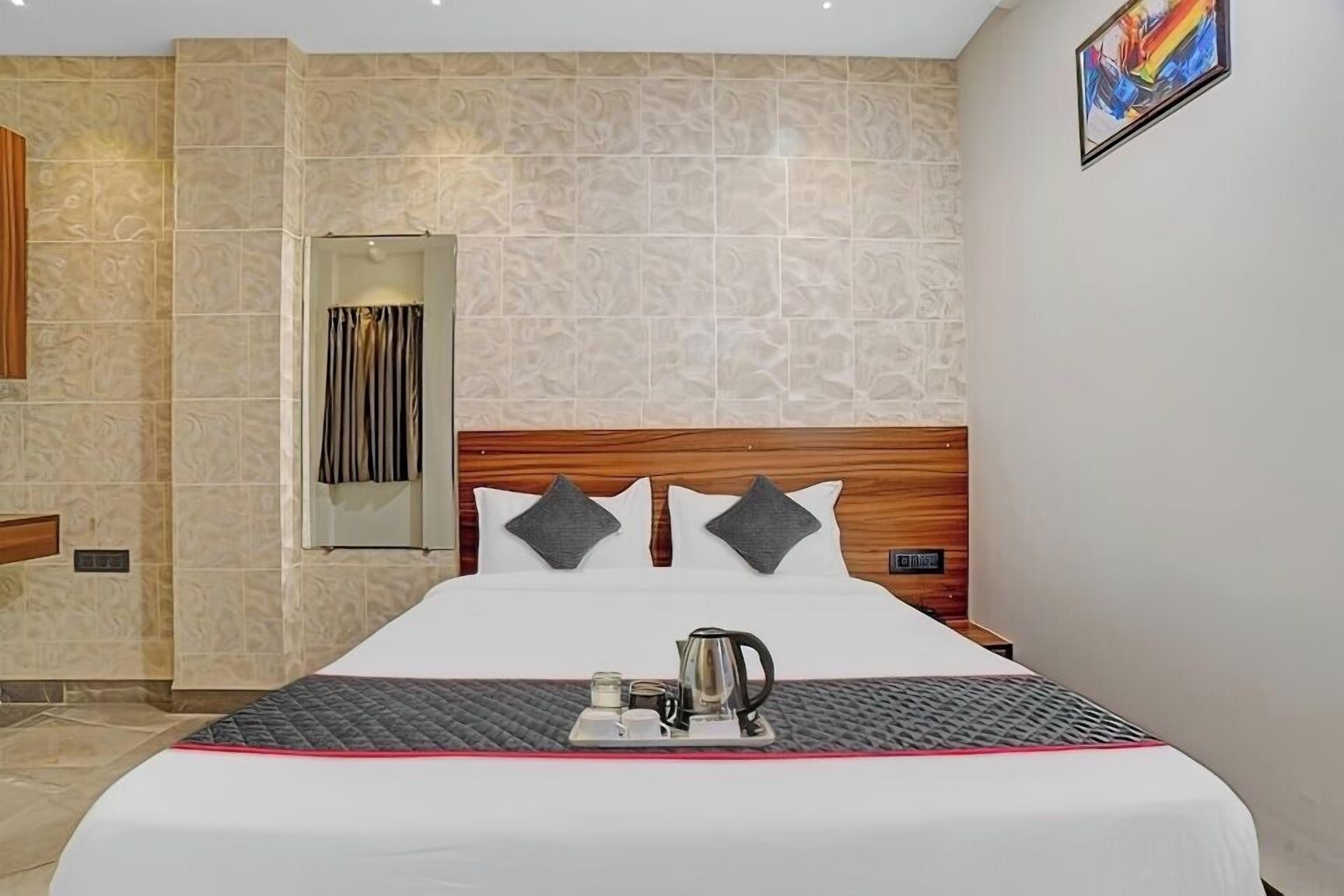 A hotel room with a large bed, a wooden headboard, two pillows, a tray with a kettle and cups, and a mirror on the wall.