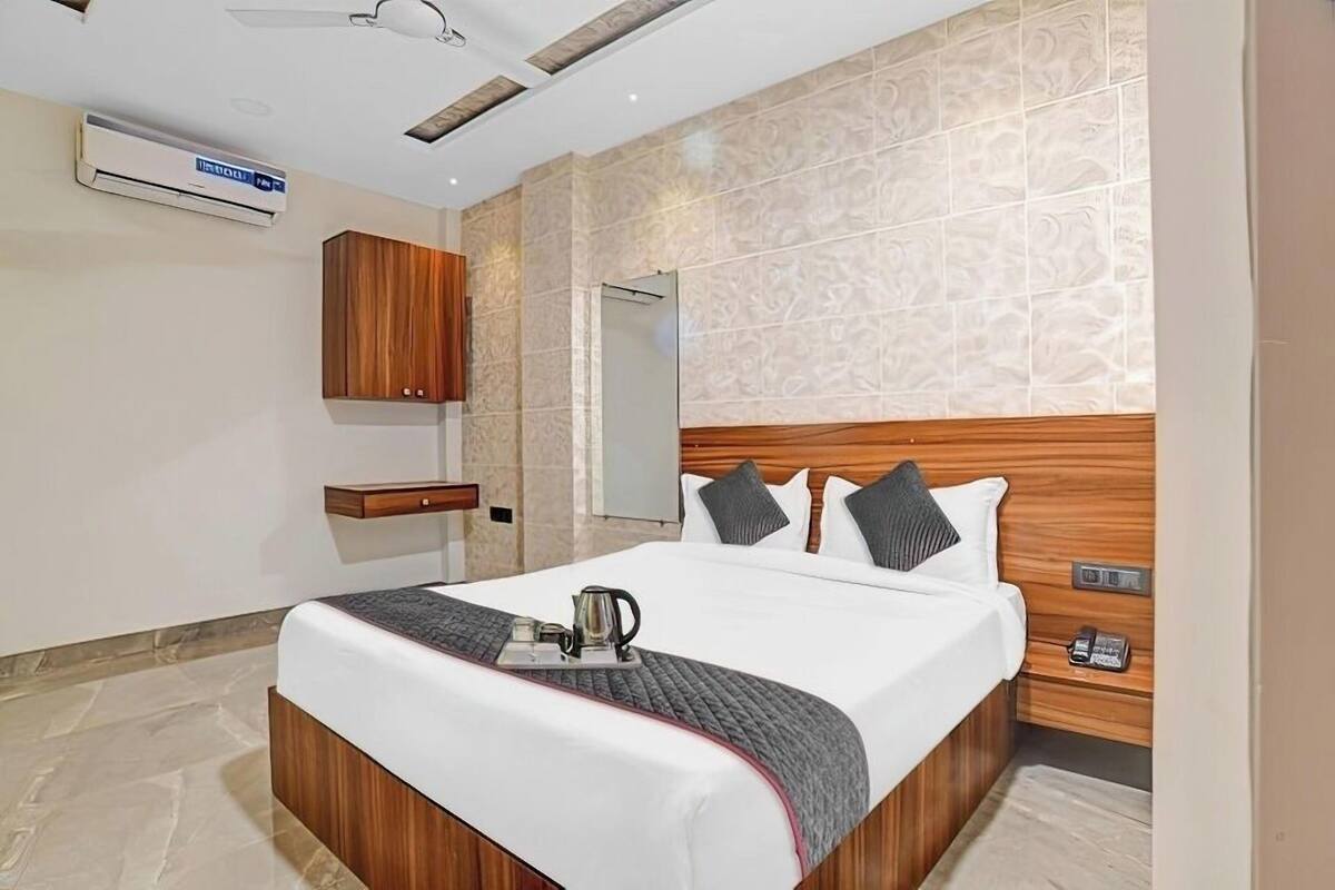 A modern hotel room with a large bed, wooden headboard, a bedside table with a lamp, and a built-in air conditioning unit.