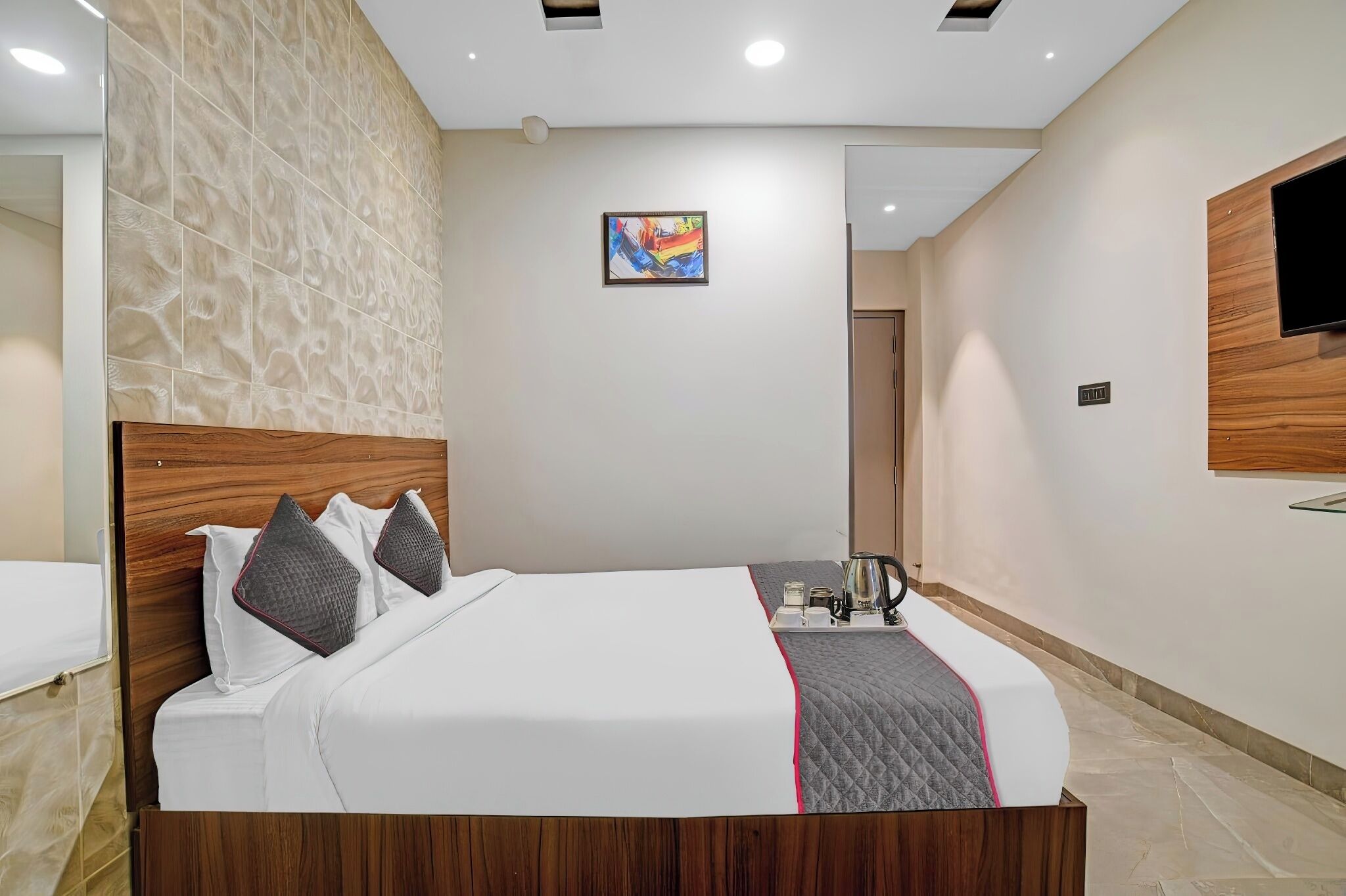 A modern hotel room with a large bed, a wooden headboard, a flat-screen TV mounted on the wall, and a painting above the bed.