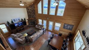 A spacious living room with a stone fireplace, large windows, and a high ceiling.