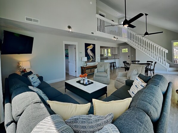 A spacious living room with a large sofa, a coffee table, and a TV. There is a staircase leading to an upper level.