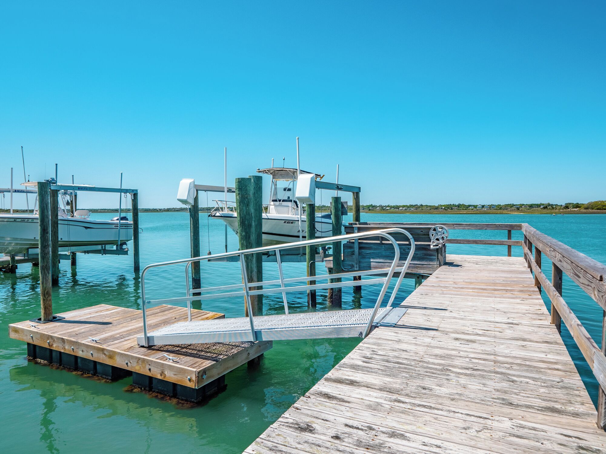 A wooden dock with a metal railing, boats docked at the pier, and clear blue water.