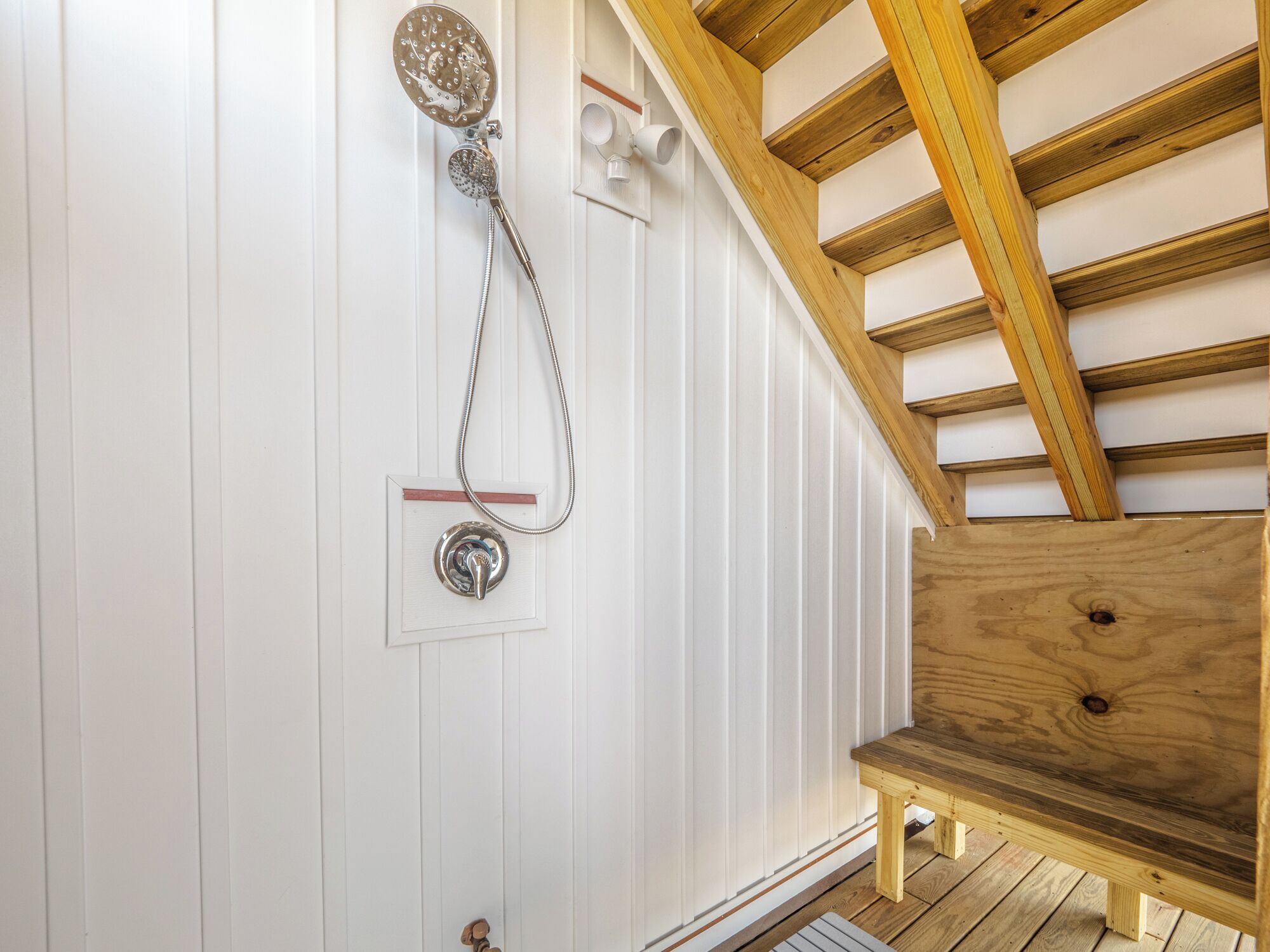 A wooden bench under a sloped ceiling with a showerhead mounted on a white wall.