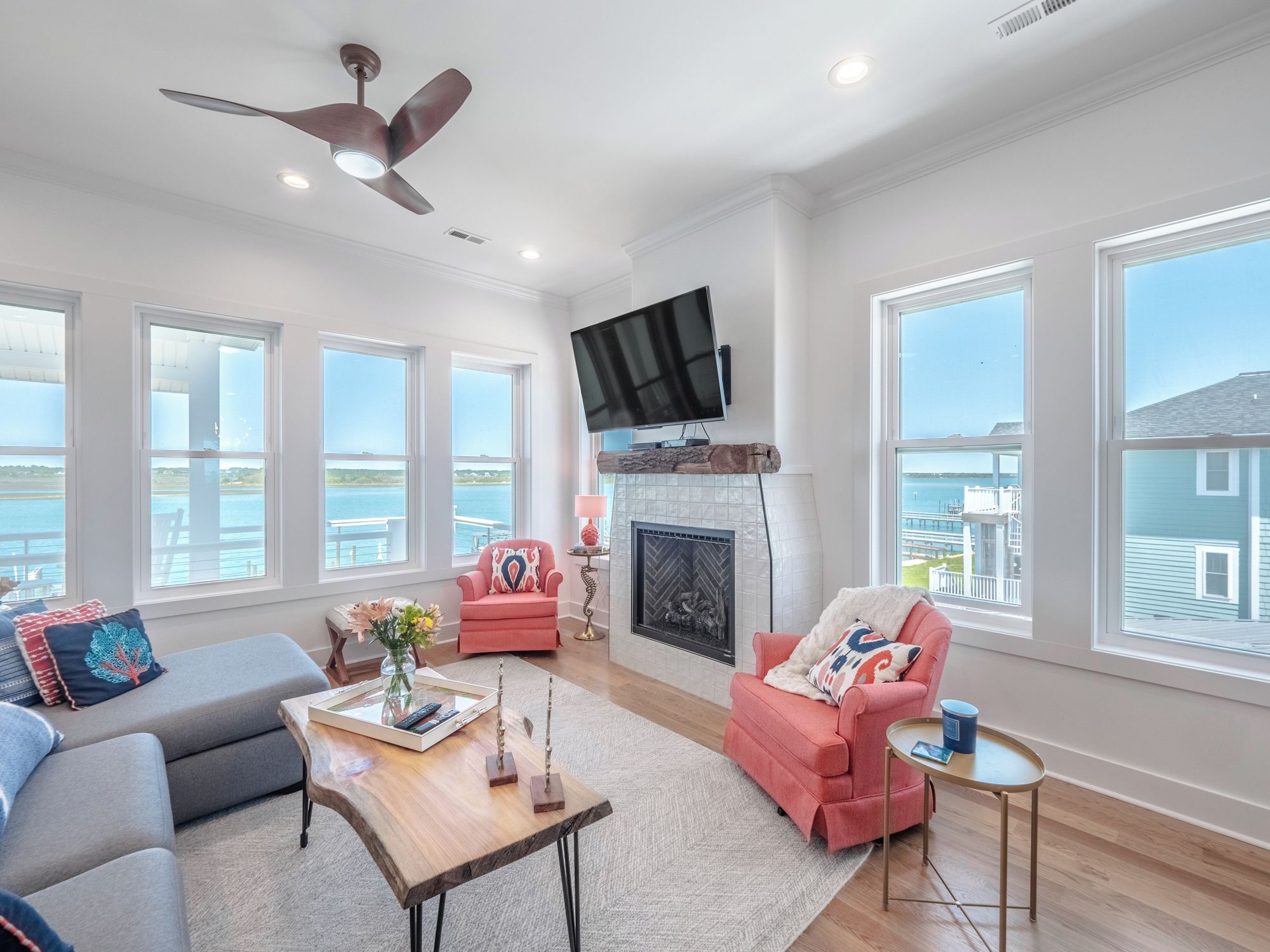 A spacious living room with a fireplace, large windows, a ceiling fan, and a flat-screen TV.