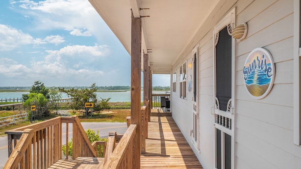 A wooden deck with a view of the water, a sign indicating a direction, and a building with a 'Sunrise' sign. - Sunrise by Topsail Realty (Holly Ridge)