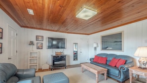 A cozy living room with a wooden ceiling, a flat-screen TV, a blue sofa with red cushions, and a coffee table. - Sunrise by Topsail Realty (Holly Ridge)