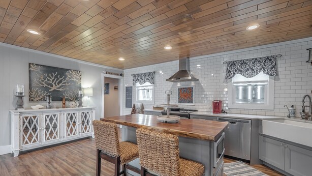 A modern kitchen with a wooden ceiling, white tiled backsplash, and a central island with bar stools. - Barefoot Bungalow by Carolina Retreats (Kure Beach)