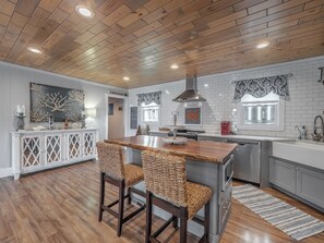A modern kitchen with a wooden ceiling, white tiled backsplash, and a central island with bar stools. - Barefoot Bungalow by Carolina Retreats (Kure Beach)