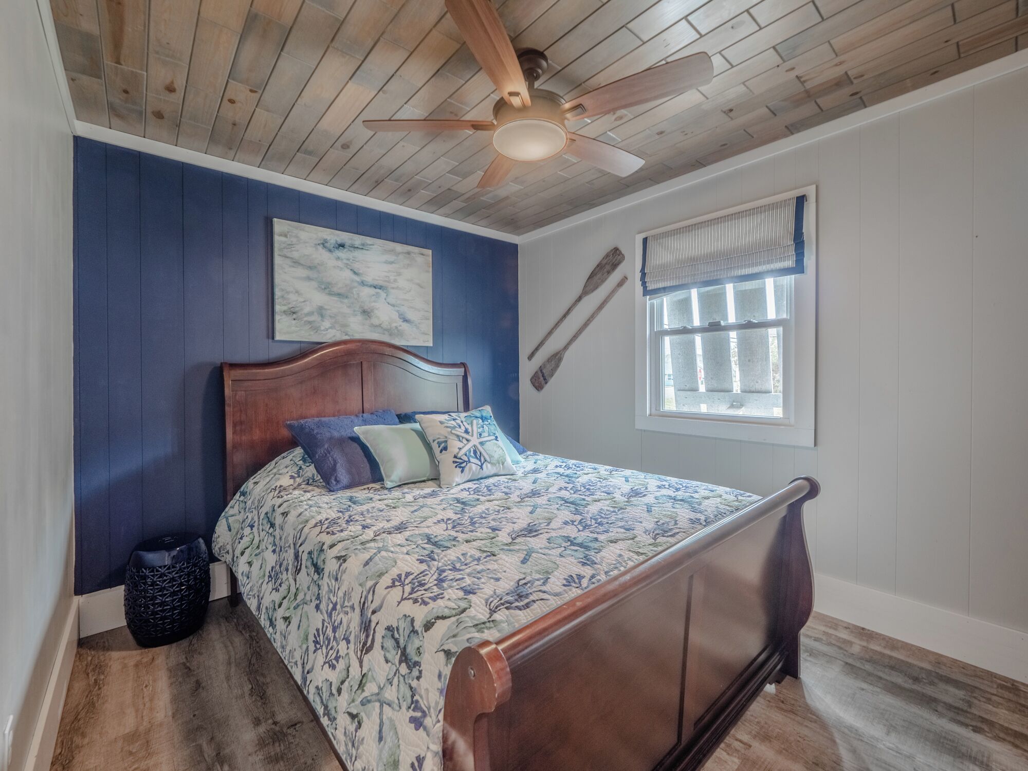 A bedroom with a wooden ceiling fan, a bed with floral bedding, a blue accent wall, and a window with a view.