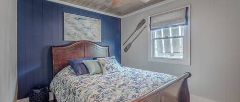 A bedroom with a wooden ceiling fan, a bed with floral bedding, a blue accent wall, and a window with a view.