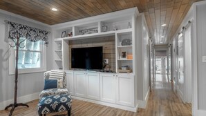 A spacious living area with a large TV, wooden flooring, and white cabinetry. - Barefoot Bungalow by Carolina Retreats (Kure Beach)
