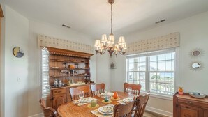 A dining room with a wooden table set for six, surrounded by chairs, a chandelier, and a window with a view. - Perfectly Tangy by Topsail Realty (Holly Ridge)