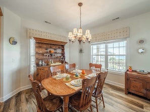 A dining room with a wooden table set for six, surrounded by chairs, a chandelier, and a window with a view.