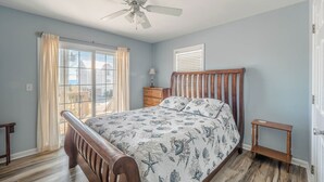 A bedroom with a bed, a ceiling fan, a window with curtains, a nightstand, and a door. - Perfectly Tangy by Topsail Realty (Holly Ridge)