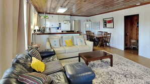 Een gezellige woonkamer met een leren bank, een houten bijzettafeltje en een vloerkleed met een patroon. Er is een keuken met witte kastjes en een eettafel met stoelen.