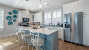 A modern kitchen with white cabinets, a stainless steel refrigerator, and a blue island with bar stools. - Serenity By the Sea by Carolina Retreats (Kure Beach)