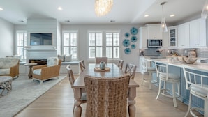 A spacious living room with a dining area, kitchen, and a large window with shutters. - Serenity By the Sea by Carolina Retreats (Kure Beach)