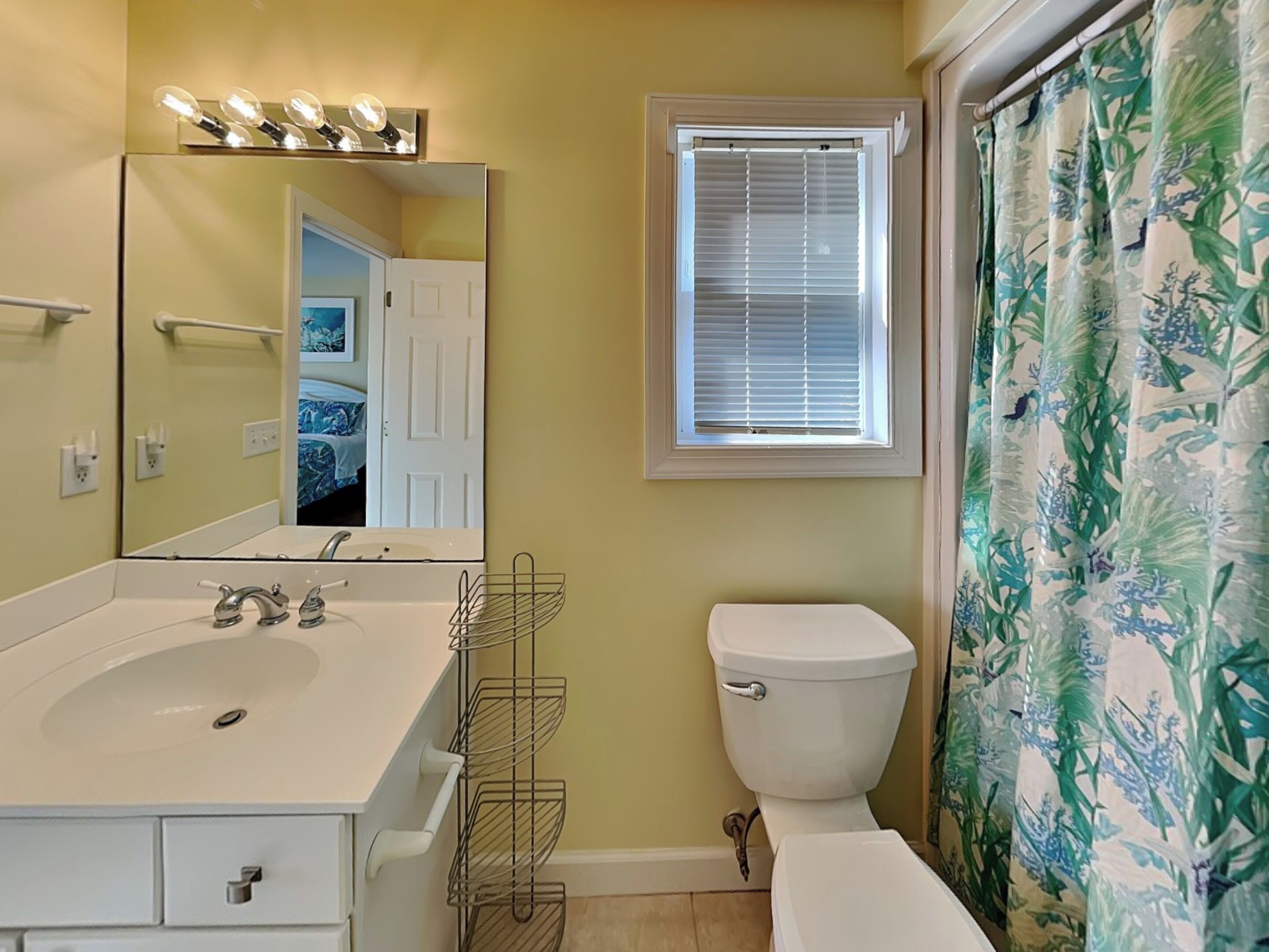 A bathroom with a white sink, mirror, and toilet. There is a window with blinds and a shower curtain with a floral pattern.